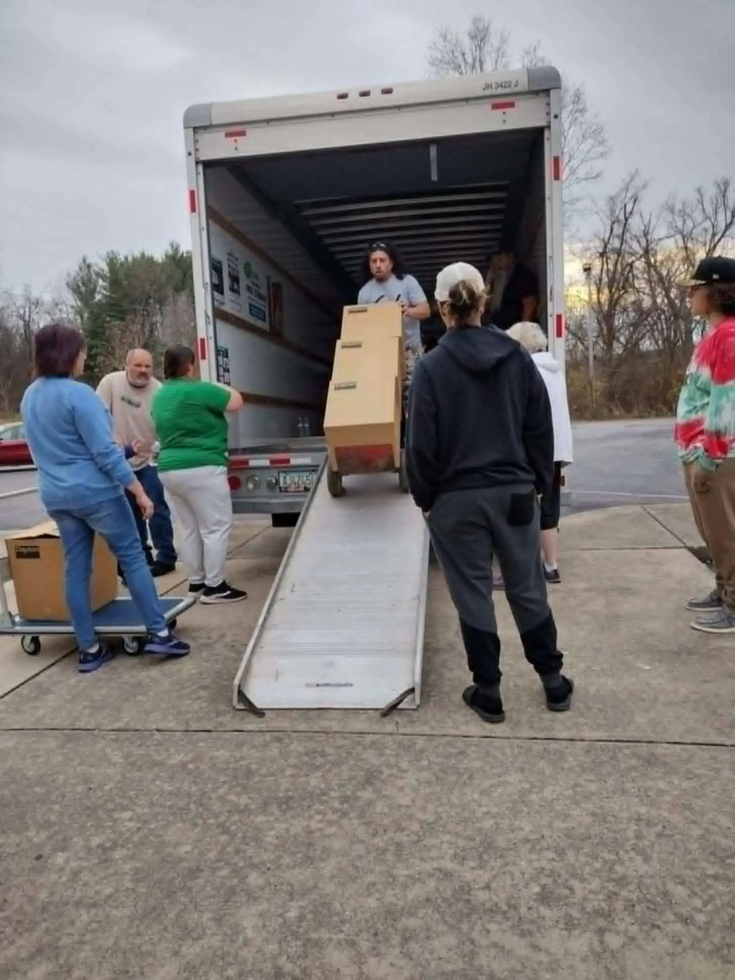 Volunteers gathered at the open back of a box truck, moving cartons down a loading ramp on hand trucks.