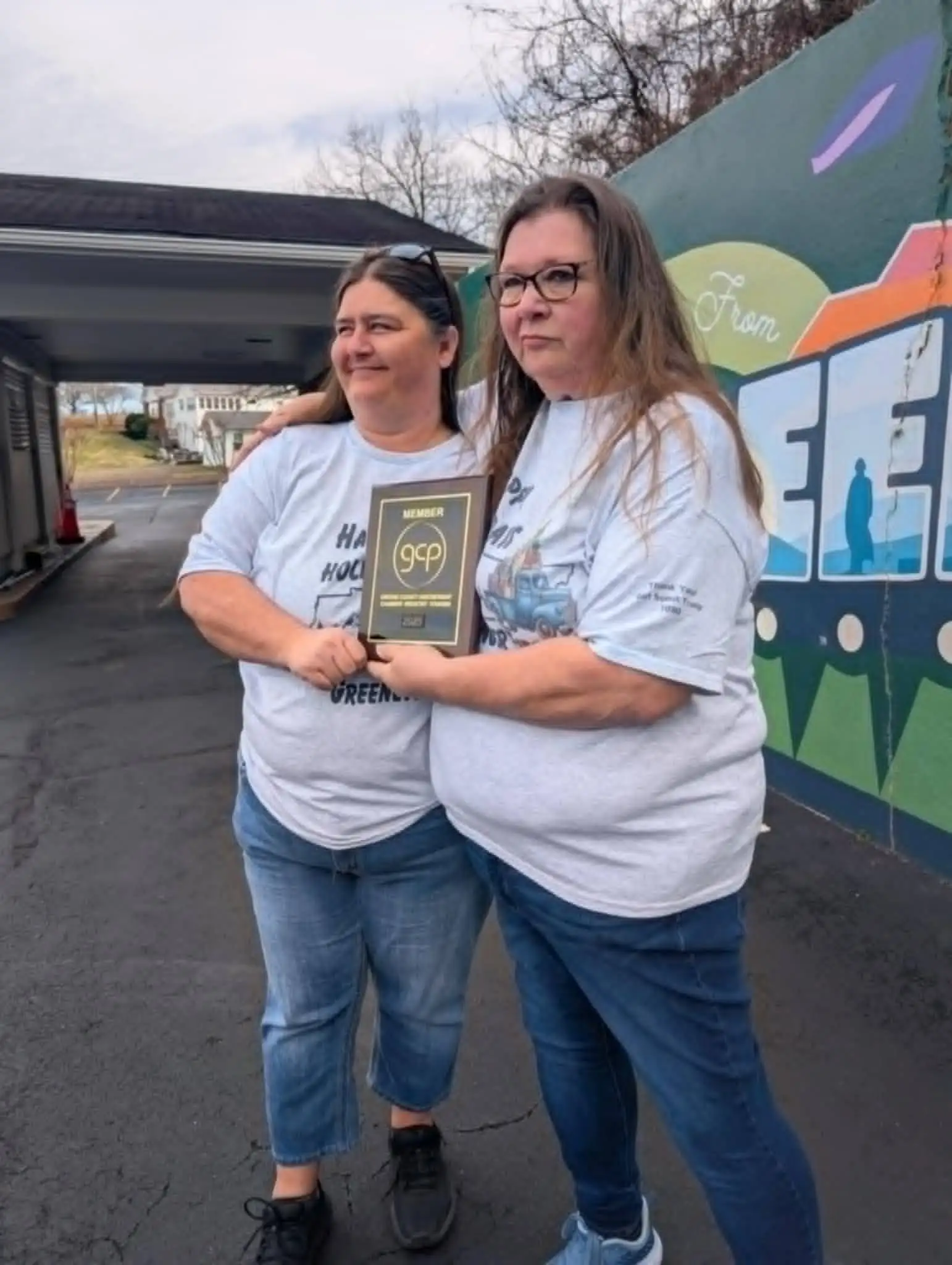 Two Hunger First volunteers in matching t-shirts holding a Greene County Partnership member plaque in front of the “Greetings from Greeneville” mural.