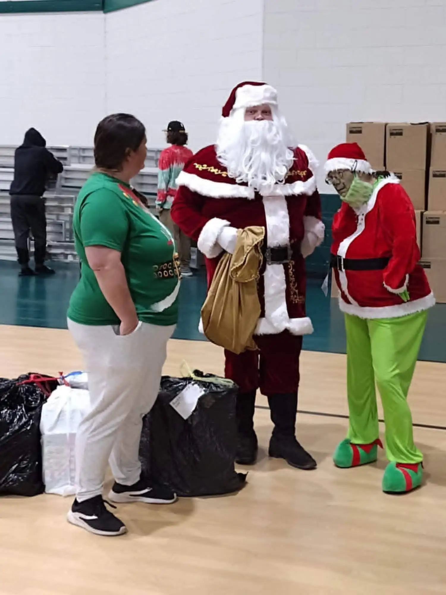 Santa and the Grinch standing with a volunteer in a gym, beside bagged donations and stacked boxes.